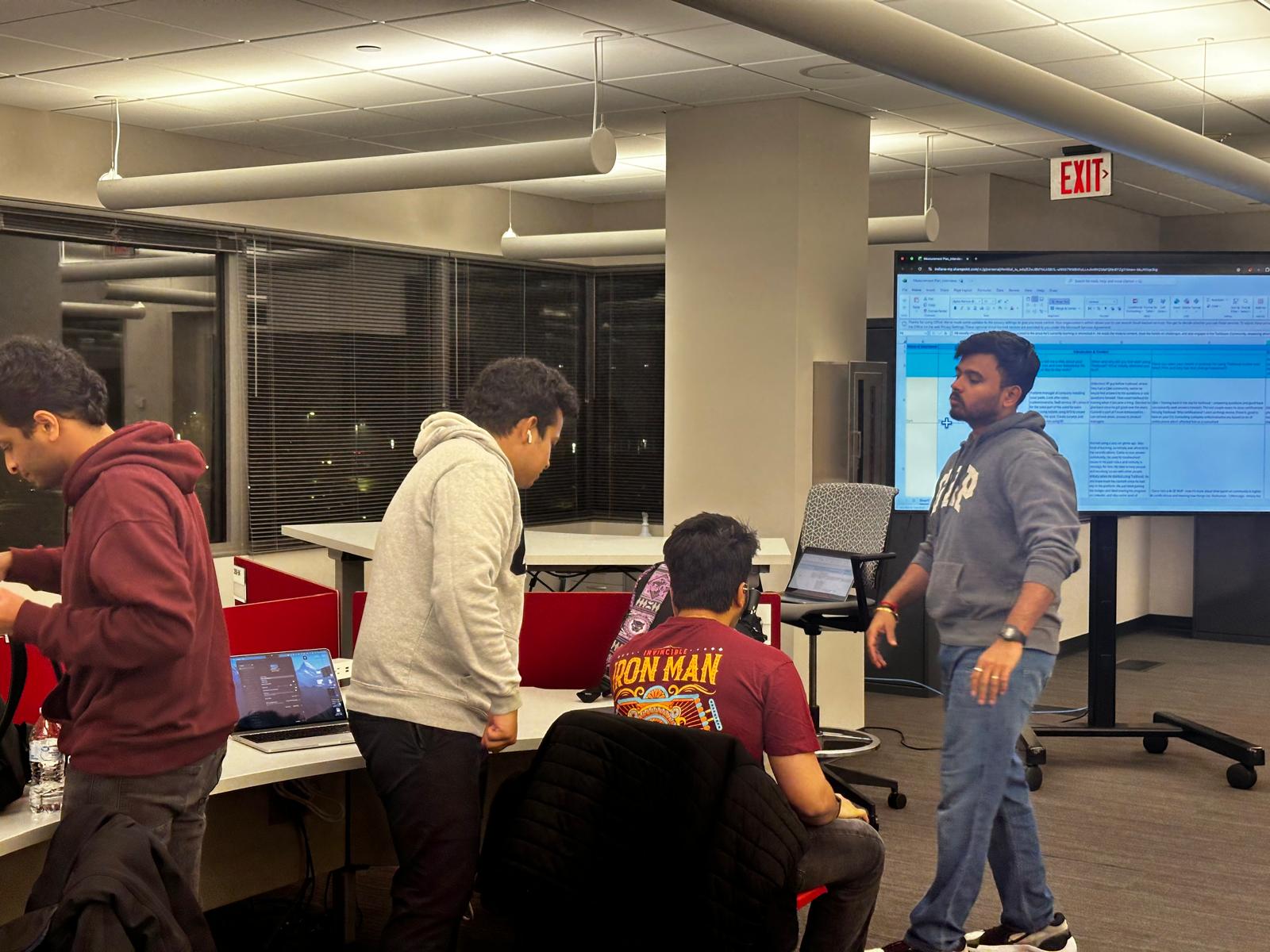 Four teammates working in an office during the ideation session, gathered around laptops while one stands beside a screen showing the interview measurement plan spreadsheet.